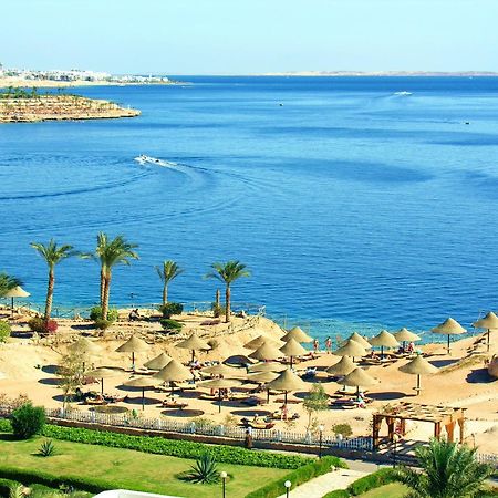 V Hotel Sharm El Shiekh Esterno foto The photo depicts a beautiful coastal scene featuring a serene blue sea and a sandy beach. There are several thatched umbrellas set up along the beach, suggesting a relaxing atmosphere for sunbathing or lounging. Palms are scattered throughout the ar