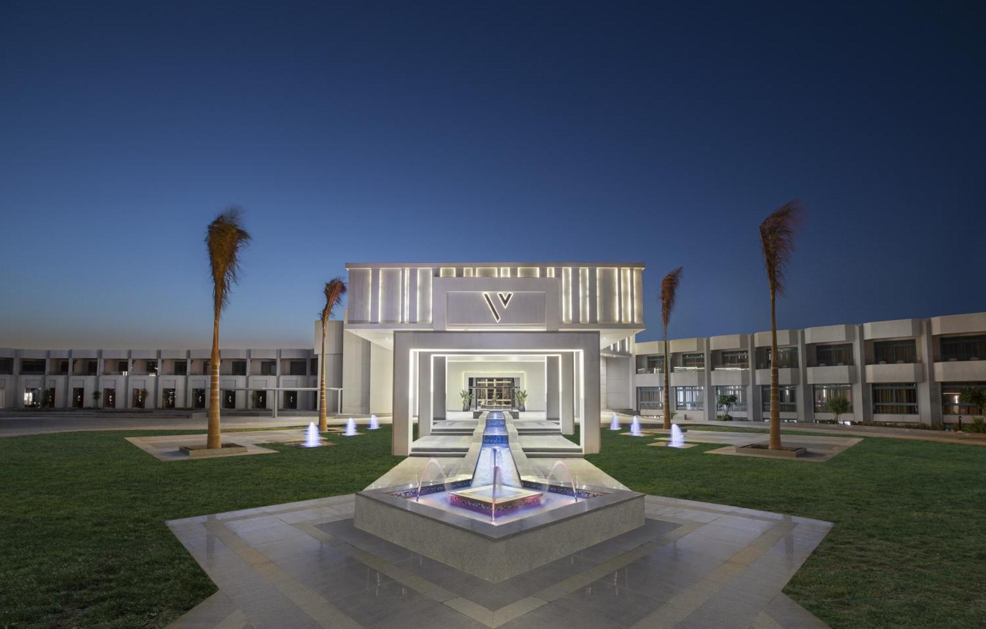 V Hotel Sharm El Shiekh Esterno foto The photo shows a modern architectural building illuminated at dusk. The structure features a central entrance framed by illuminated elements and has a sleek, contemporary design. In front of the building, there is a water feature with a fountain, wh
