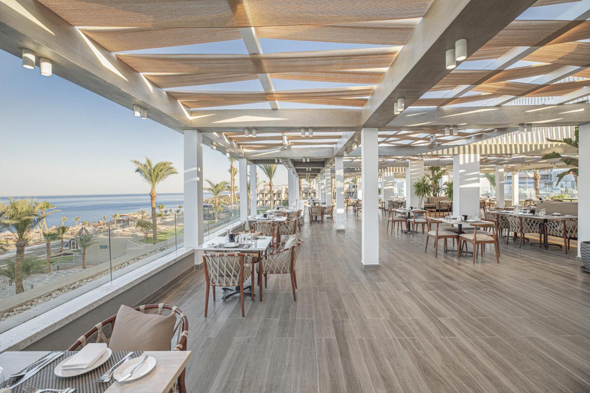 V Hotel Sharm El Shiekh Esterno foto The photo shows a stylish restaurant interior with a modern design. It features large windows that provide a panoramic view of the beach or ocean beyond. The space is bright and airy, enhanced by wooden beams on the ceiling and ample natural light. T