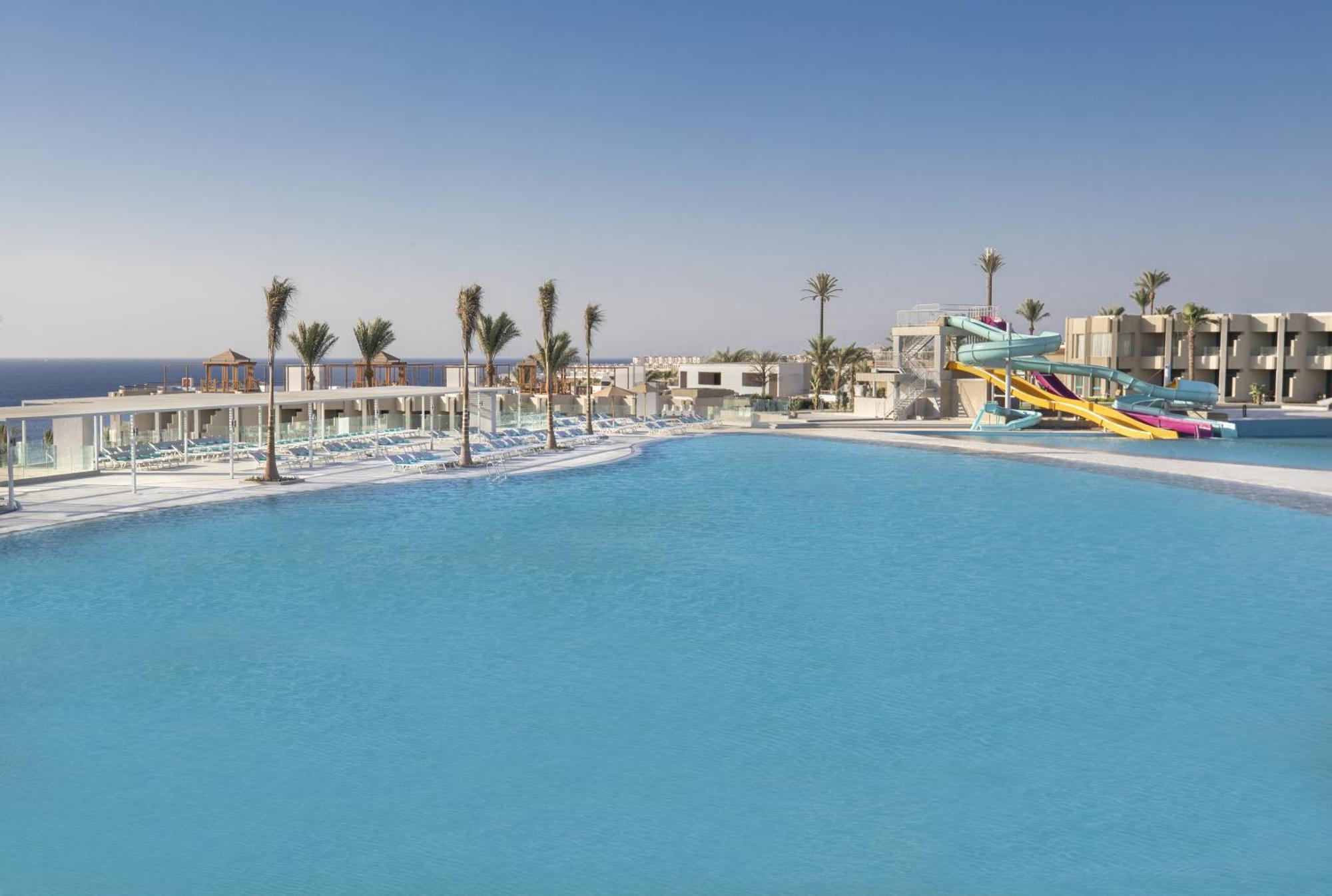 V Hotel Sharm El Shiekh Esterno foto The photo shows a sunny outdoor pool area with clear blue water. In the foreground, there is a large swimming pool, and in the background, several palm trees line the edges. To the right, a colorful water slide is visible, suggesting a fun recreation