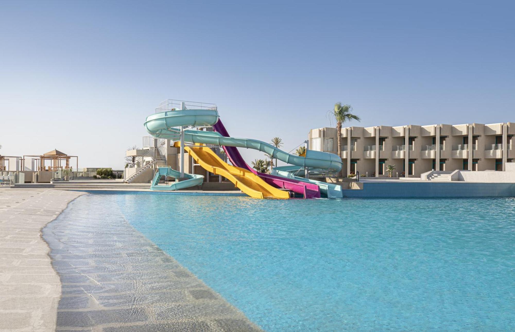 V Hotel Sharm El Shiekh Esterno foto The photo depicts a vibrant swimming pool area featuring two large water slides. One slide is turquoise and the other is yellow with purple accents. The pool is expansive, with clear blue water, and there are palm trees in the background, suggesting 
