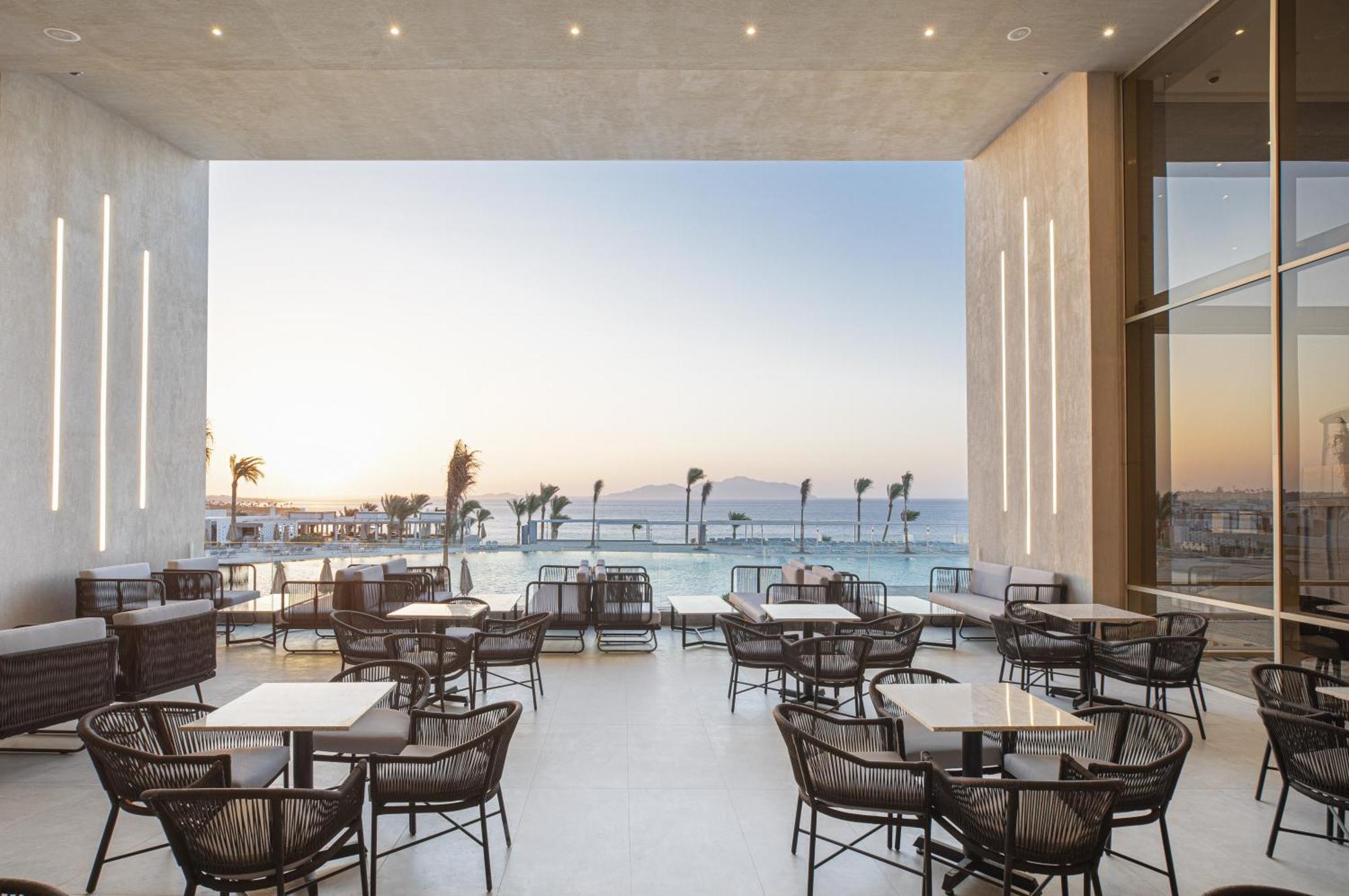V Hotel Sharm El Shiekh Esterno foto The photo depicts a modern indoor dining area with several wicker chairs and tables arranged neatly. Large glass panels create an open space that offers a scenic view of the sea and a sunset in the background, where gentle colors of orange and blue b
