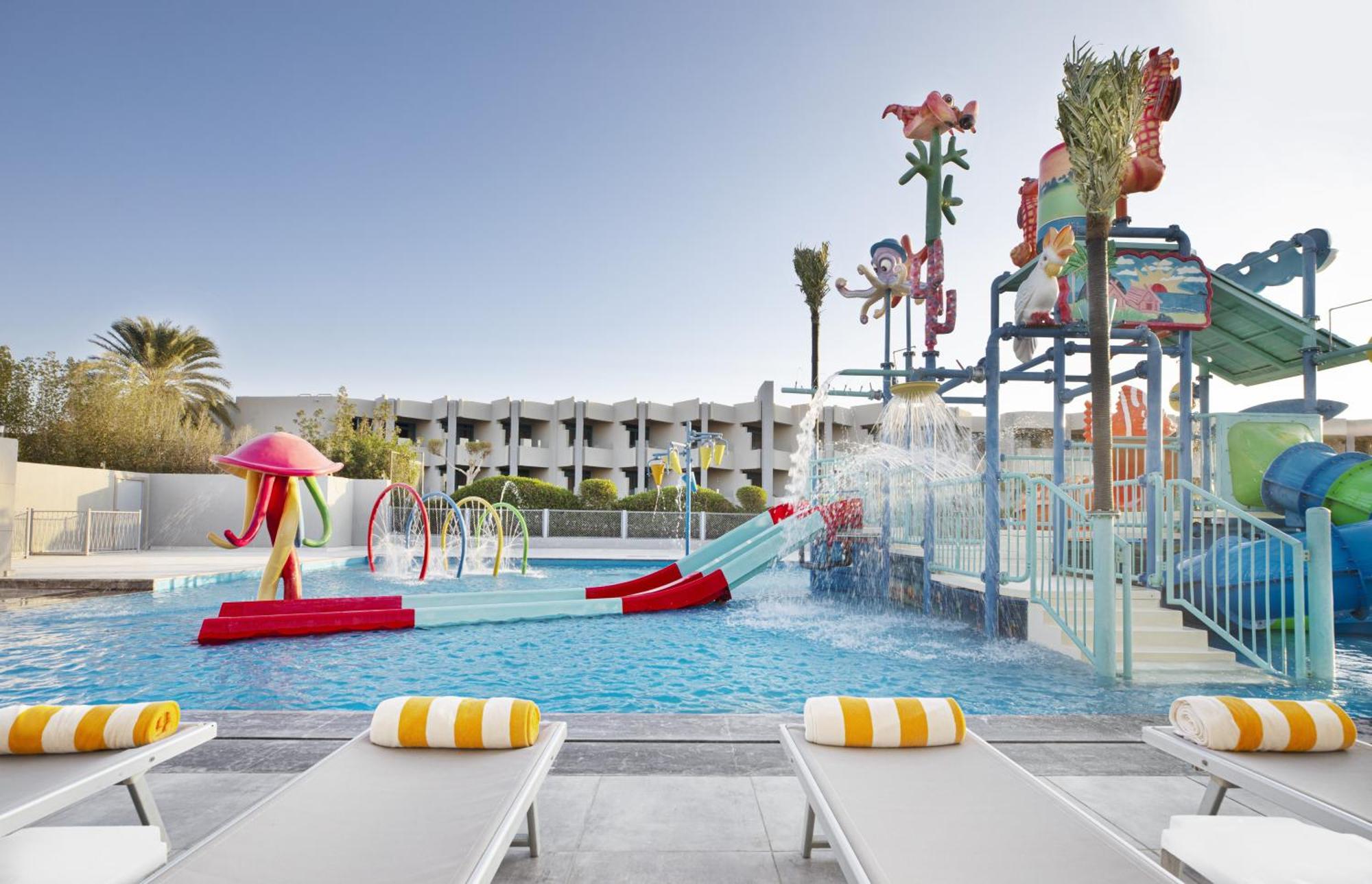 V Hotel Sharm El Shiekh Esterno foto The photo shows a vibrant and colorful water park area. There is a large swimming pool with various water features, including slides and spray elements. The pool area is decorated with bright, playful designs, such as sea creatures and tropical theme