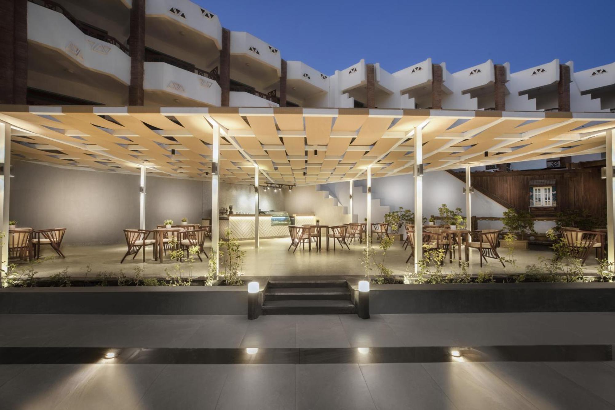 V Hotel Sharm El Shiekh Esterno foto The photo depicts a modern outdoor dining area or café setting. The space features a structured canopy made of grid-like panels, providing shade. Below the canopy, there are several wooden tables and chairs arranged neatly. The area is well-lit, with