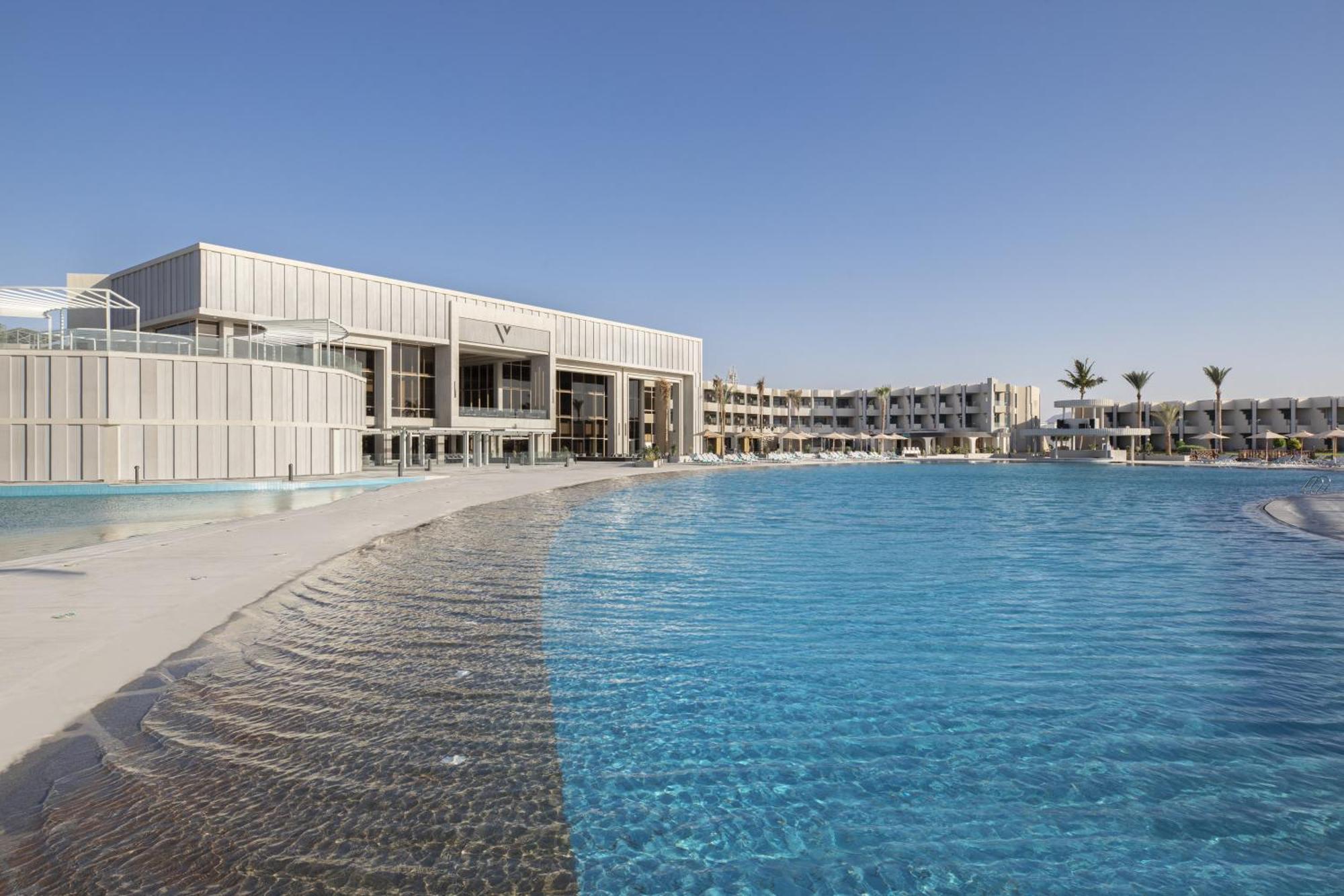 V Hotel Sharm El Shiekh Esterno foto The photo shows a modern hotel or resort building situated near a large swimming pool. The pool has a gentle, wavy edge and is filled with clear blue water. In the background, there are multiple buildings that appear to be part of the same resort com