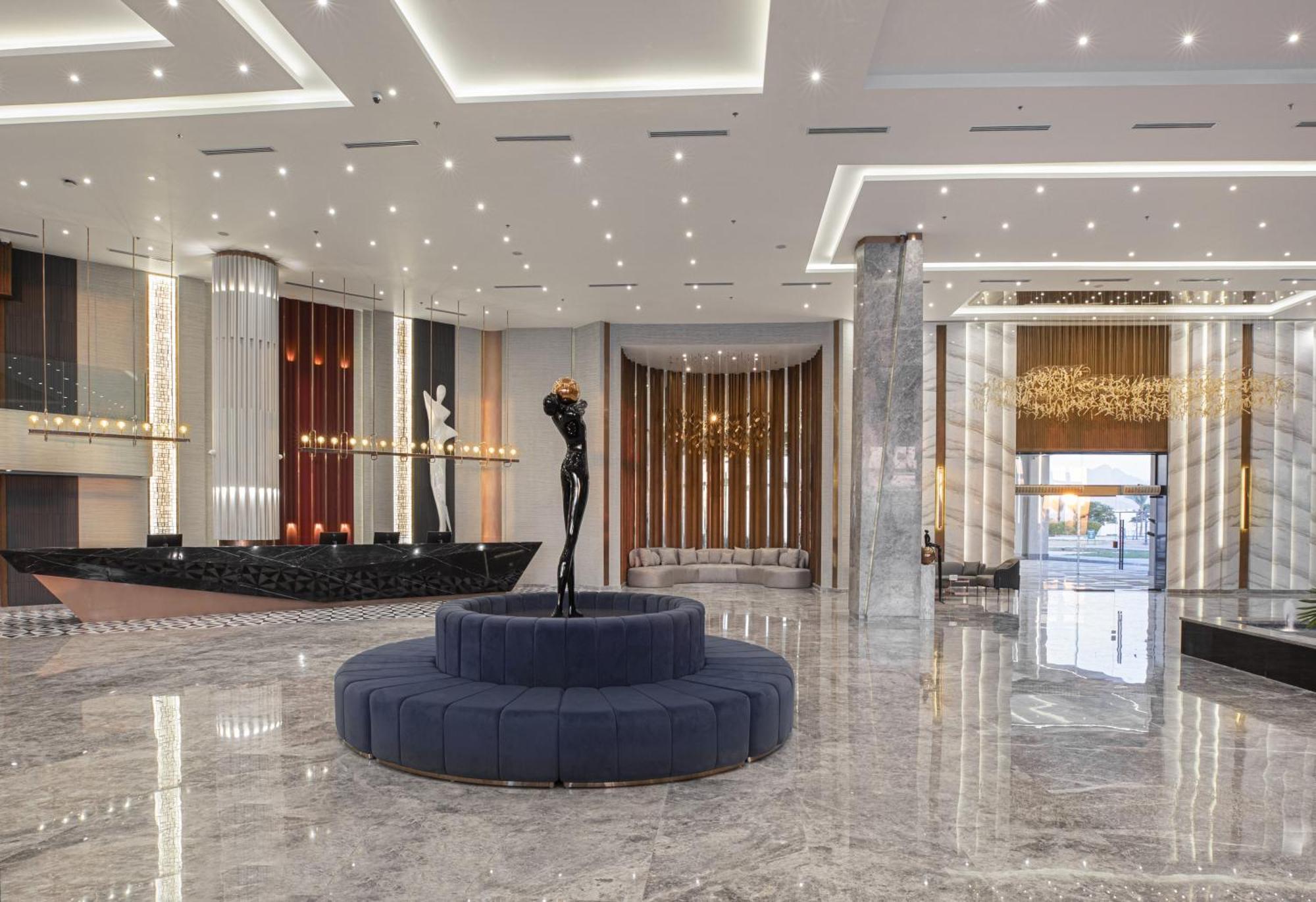 V Hotel Sharm El Shiekh Esterno foto The photo displays a modern hotel lobby with a sleek and elegant design. The flooring is made of polished marble, reflecting the ambient light. In the center, there is a circular blue velvet seating area surrounded by a minimalist black sculpture. Th