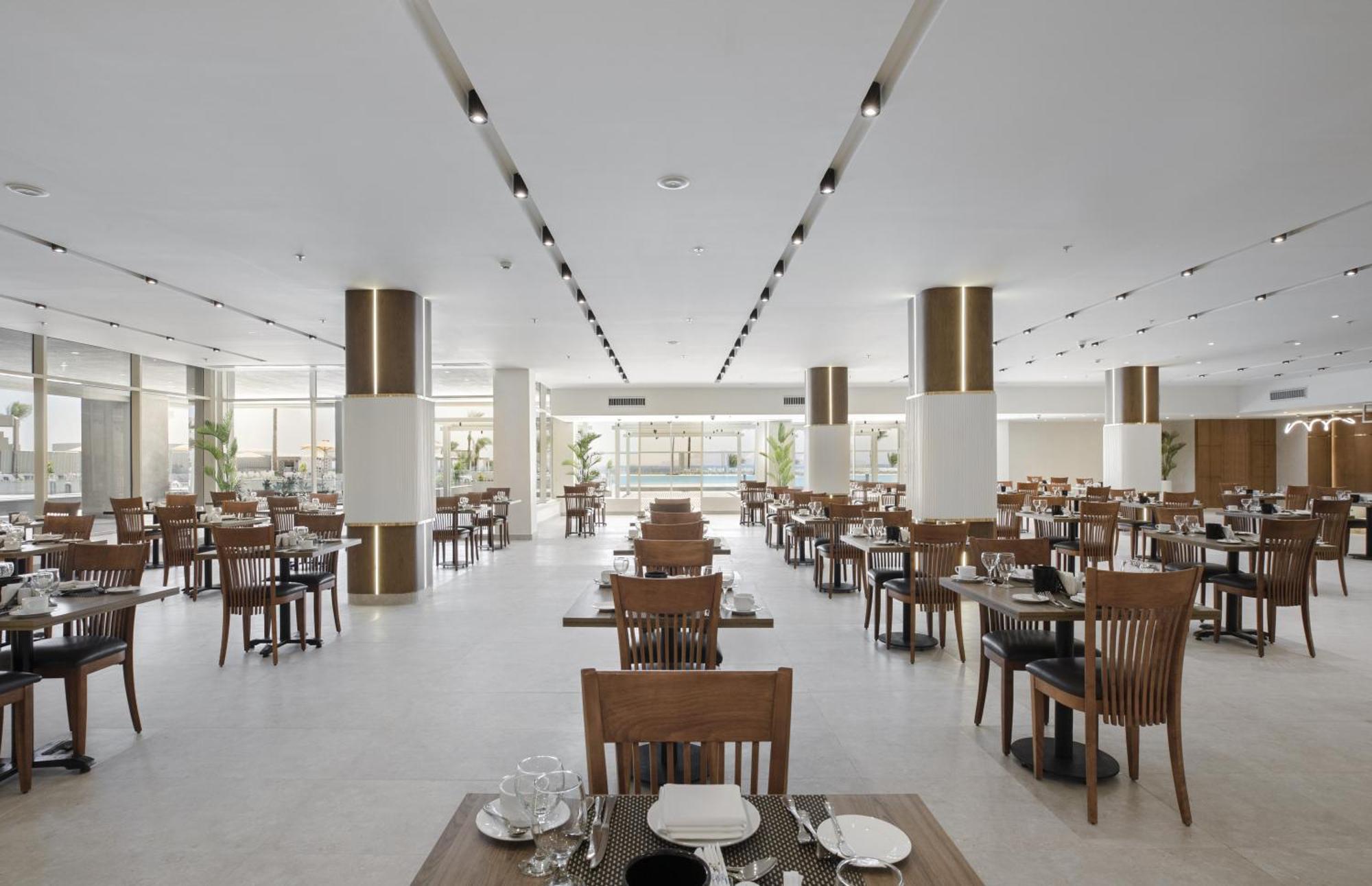 V Hotel Sharm El Shiekh Esterno foto The photo shows a spacious and modern restaurant interior. It features neatly arranged wooden tables with chairs and white table settings, including plates and cutlery. The design is minimalist with light-colored walls and polished flooring. There ar