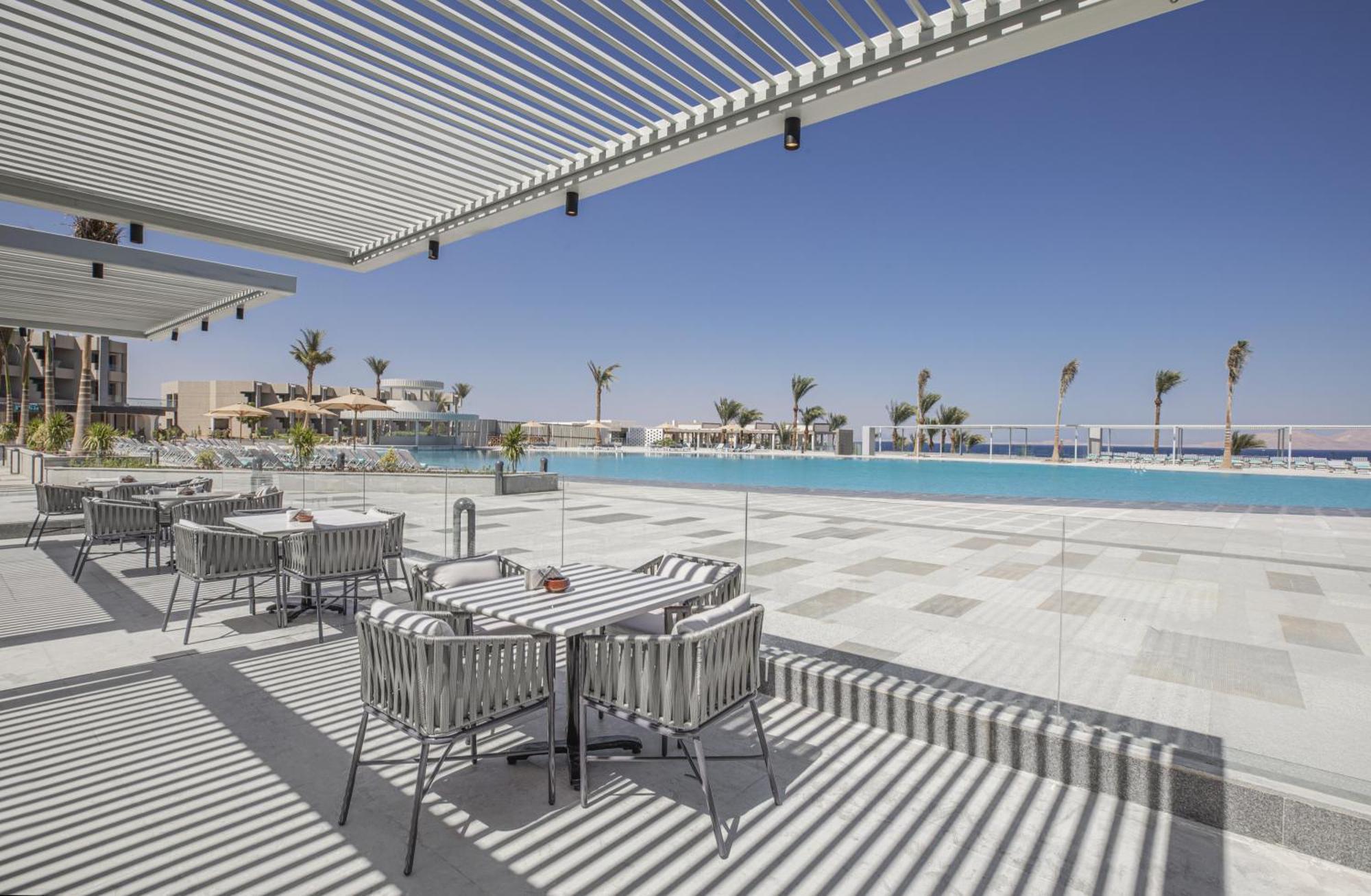 V Hotel Sharm El Shiekh Esterno foto The photo depicts a modern outdoor setting, likely at a resort or hotel. In the foreground, there are several stylish dining tables and chairs, arranged neatly under a shaded area created by slatted overhead structures. The background features a larg