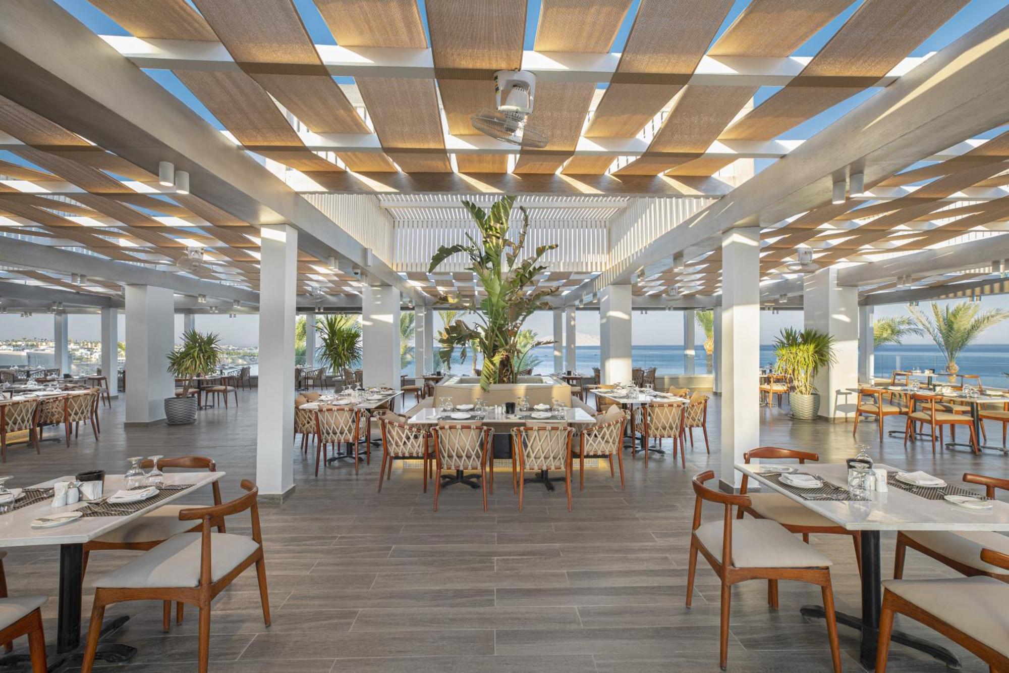 V Hotel Sharm El Shiekh Esterno foto The photo shows a spacious and modern restaurant interior. It features a bright and airy atmosphere with large windows letting in natural light. The ceiling has a series of wooden slats, creating a stylish pattern and providing shade. There's a centr