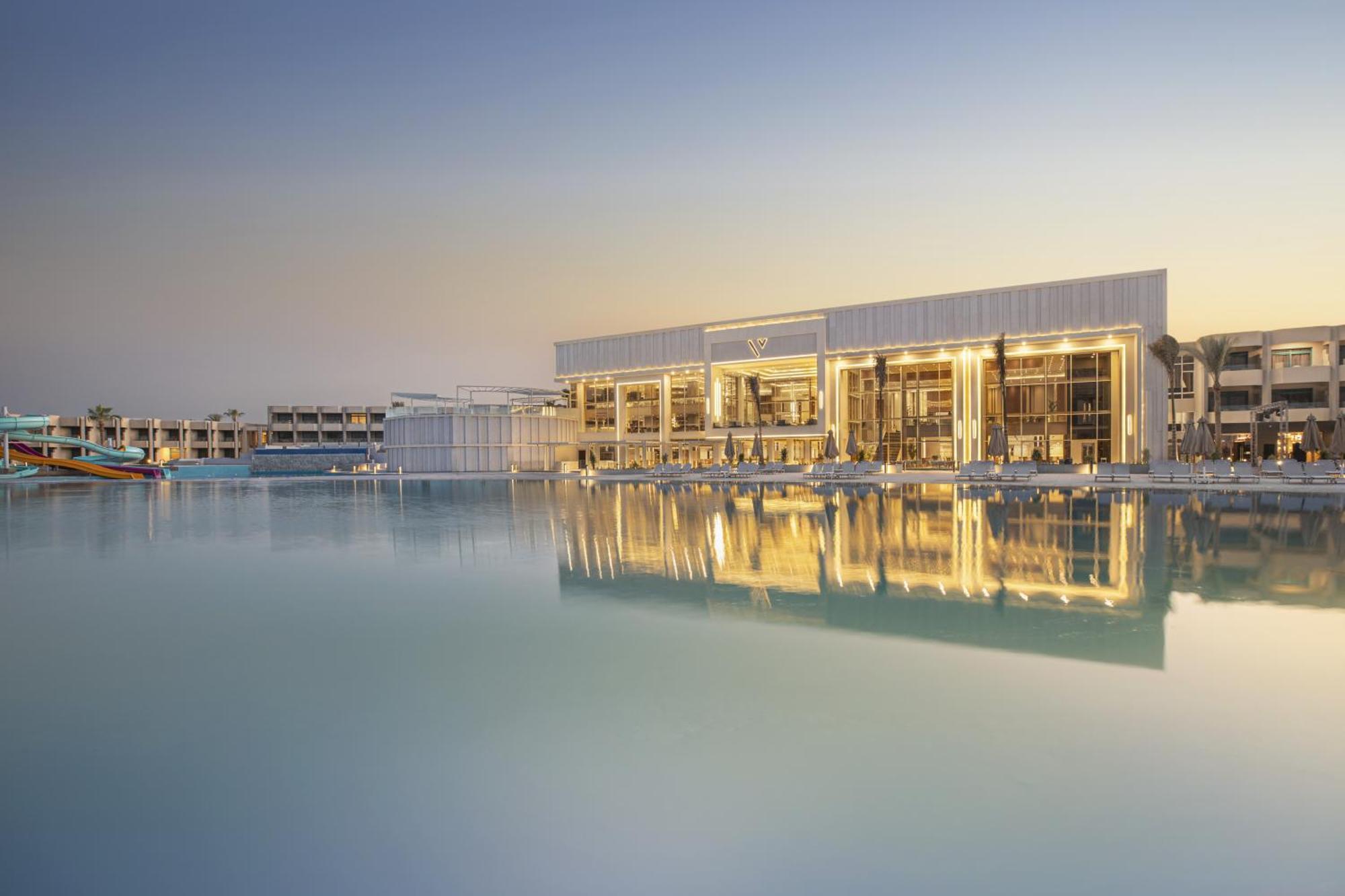 V Hotel Sharm El Shiekh Esterno foto The photo depicts a modern building situated by a calm body of water, likely a lake or lagoon. The structure features a sleek design with large glass windows that reflect the water and the surrounding environment. The setting appears tranquil, possib