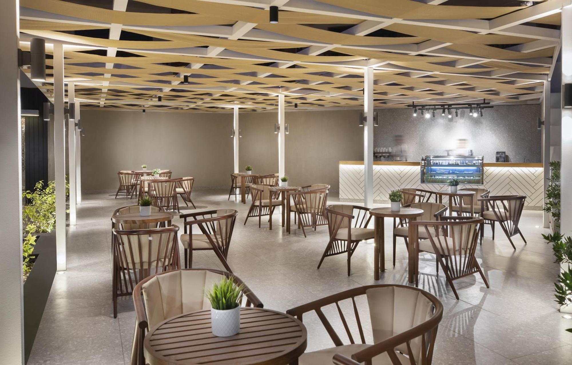 V Hotel Sharm El Shiekh Esterno foto The photo shows a modern indoor cafe or restaurant space. It features a series of wooden tables and chairs arranged throughout the area, with several plants placed on the tables. The ceiling has a distinctive geometric design with wooden beams, creat