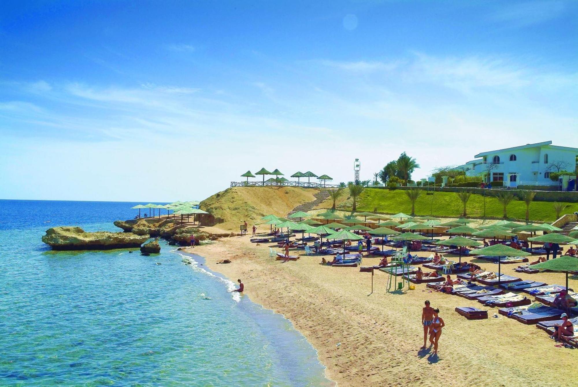 V Hotel Sharm El Shiekh Esterno foto The photo depicts a picturesque beach scene. There is a sandy shoreline with clear blue water gently lapping at the shore. Green umbrellas and lounge chairs are neatly arranged on the sand, providing shade for relaxation. In the background, there is 