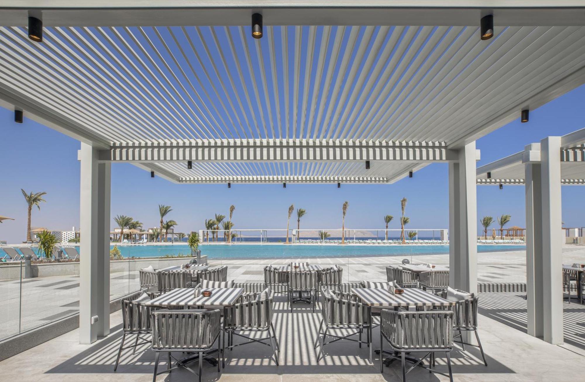 V Hotel Sharm El Shiekh Esterno foto The photo shows a modern outdoor dining area with a stylish pergola overhead. Beneath the pergola are several dining tables and chairs arranged in a neat layout. The setting appears to be near a swimming pool, with a clear blue sky and palm trees vis