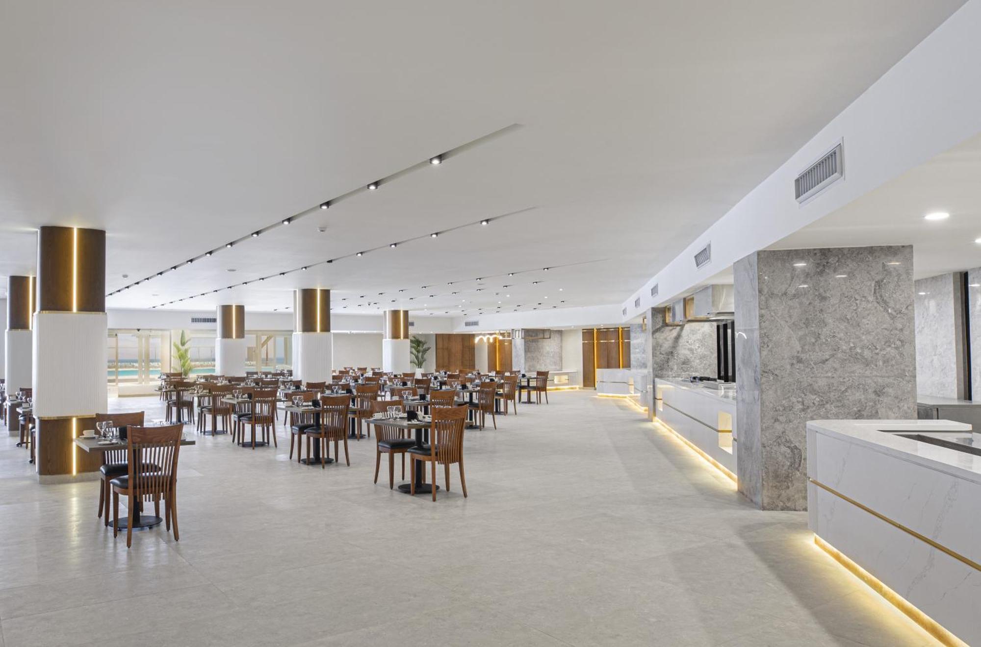 V Hotel Sharm El Shiekh Esterno foto The photo shows a modern restaurant interior. It features a spacious, well-lit environment with a clean design. The flooring is light-colored, and the walls have a minimalist aesthetic. There are several wooden tables and chairs arranged neatly throu