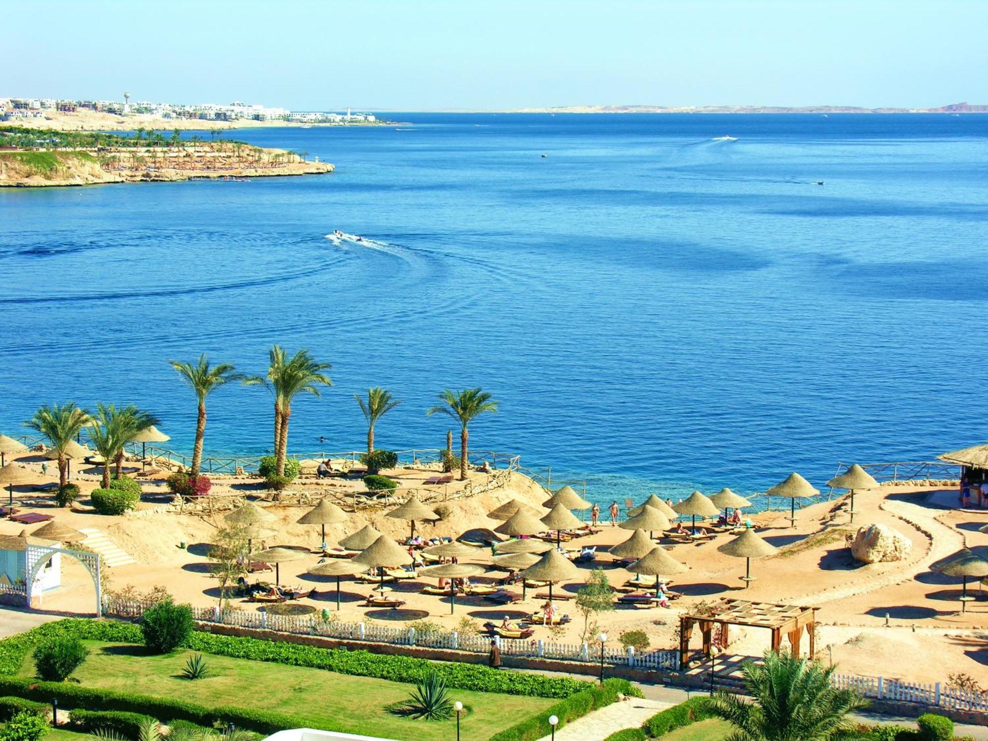 V Hotel Sharm El Shiekh Esterno foto The photo depicts a beautiful coastal scene featuring a serene blue sea and a sandy beach. There are several thatched umbrellas set up along the beach, suggesting a relaxing atmosphere for sunbathing or lounging. Palms are scattered throughout the ar